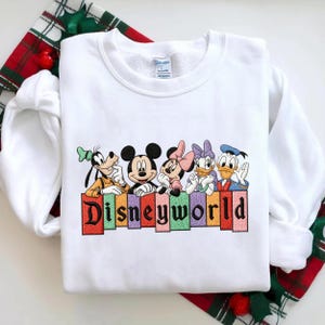 May include: White sweatshirt featuring embroidered Disney characters: Goofy, Mickey Mouse, Minnie Mouse, Daisy Duck, and Donald Duck. The word "Disneyworld" is embroidered below the characters in colorful letters. The sweatshirt is displayed on a plaid fabric.