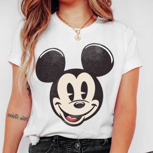 May include: White t-shirt featuring a large, smiling Mickey Mouse face in black and white. The classic cartoon character's face is centered on the shirt, with a red mouth and a happy expression. The shirt has short sleeves and a crew neck.