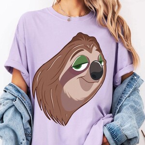 May include: A light purple t-shirt featuring a cartoon sloth face with brown fur, green eyes, and a black nose. The sloth has a relaxed expression. The shirt has rolled-up sleeves.
