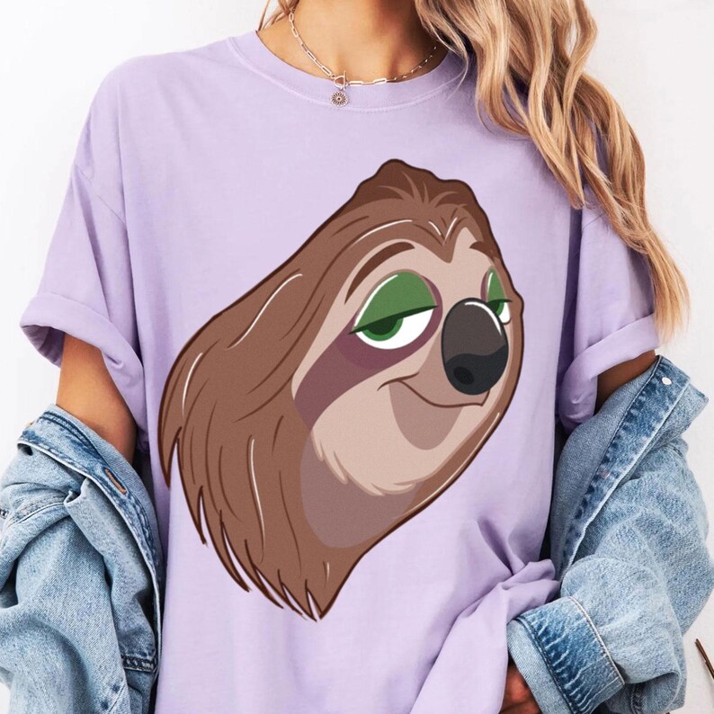 May include: A light purple t-shirt featuring a cartoon sloth face with brown fur, green eyes, and a black nose. The sloth has a relaxed expression. The shirt has rolled-up sleeves.