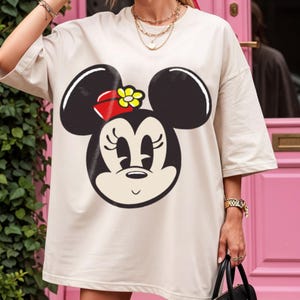 Peut inclure: T-shirt beige oversize avec un grand motif de Minnie Mouse. Le motif repr&eacute;sente le visage de Minnie en noir et blanc, avec un n&oelig;ud rouge et une fleur jaune. Le t-shirt a des manches courtes et une coupe ample.