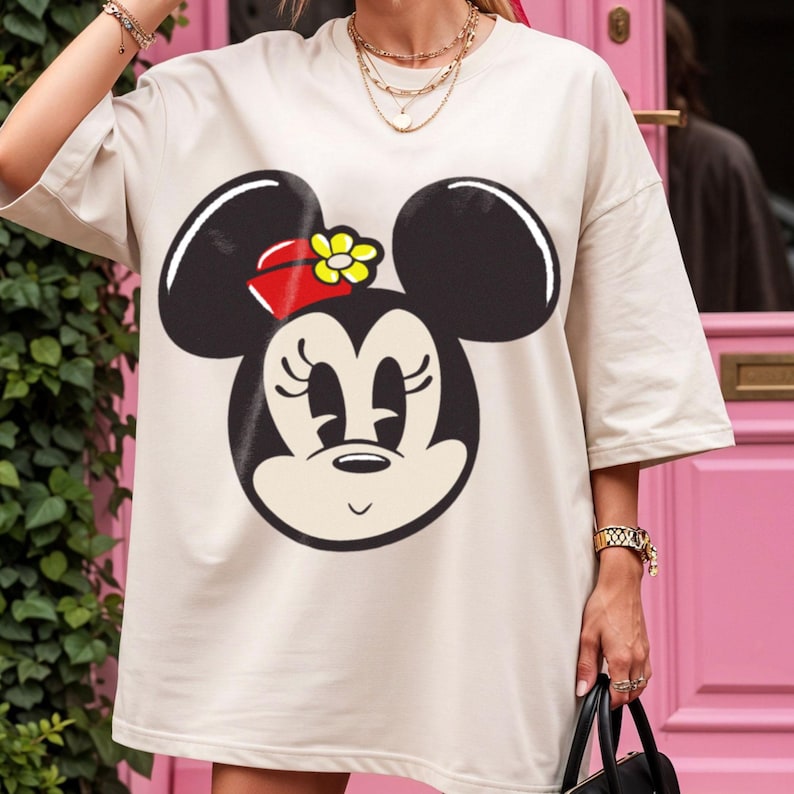 Peut inclure: T-shirt beige oversize avec un grand motif de Minnie Mouse. Le motif repr&eacute;sente le visage de Minnie en noir et blanc, avec un n&oelig;ud rouge et une fleur jaune. Le t-shirt a des manches courtes et une coupe ample.