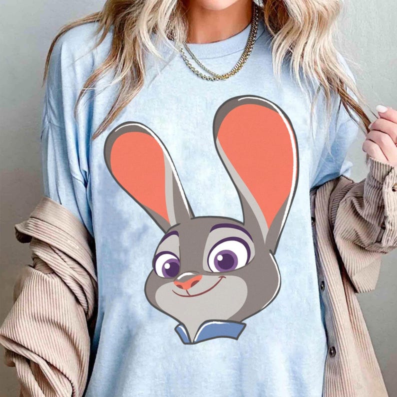 Puede incluir: Camiseta azul claro con una ilustraci&oacute;n de dibujos animados de un conejo gris sonriente con grandes orejas forradas de naranja y ojos morados. El conejo tiene un cuello azul. Una prenda informal.