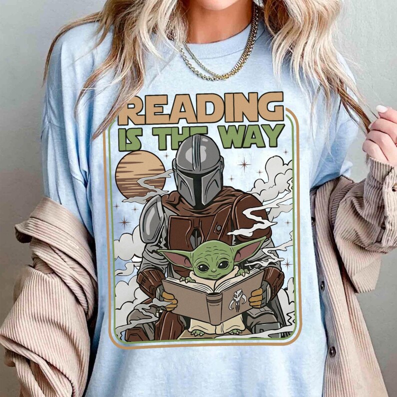 Puede incluir: Camiseta azul claro con un gr&aacute;fico de The Mandalorian y Grogu leyendo un libro. El texto "READING IS THE WAY" est&aacute; encima de los personajes. El dise&ntilde;o incluye un planeta y nubes, con un borde verde y marr&oacute;n.