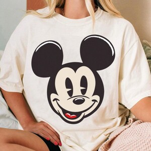 May include: Cream-colored t-shirt featuring a large graphic of Mickey Mouse's face. The design showcases Mickey's iconic black ears, smiling face, and red mouth. The shirt is a casual, comfortable style.