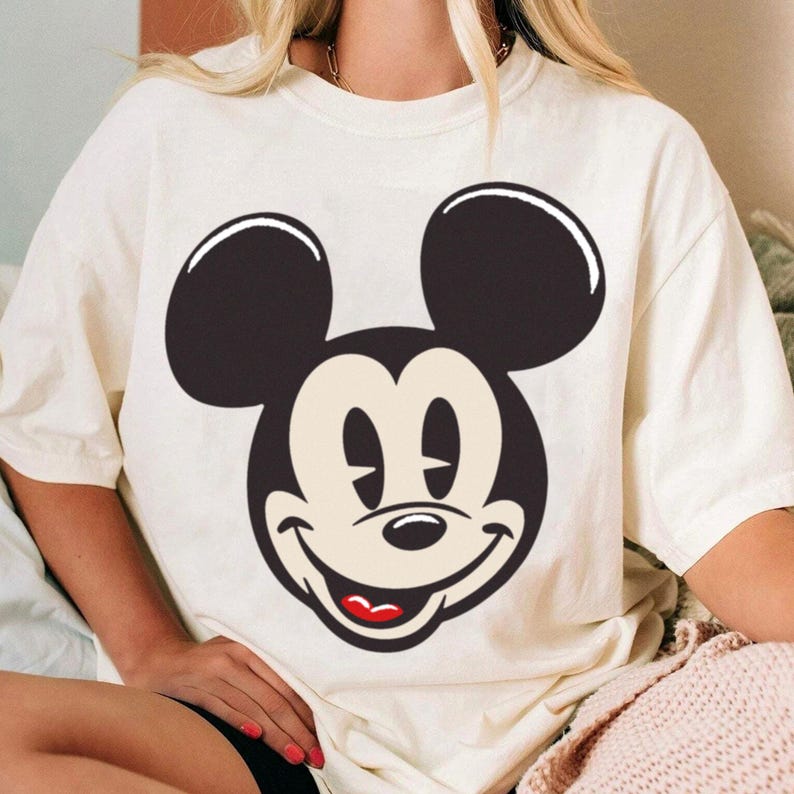 May include: Cream-colored t-shirt featuring a large graphic of Mickey Mouse's face. The design showcases Mickey's iconic black ears, smiling face, and red mouth. The shirt is a casual, comfortable style.