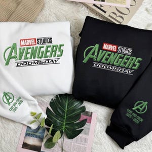 May include: White and black sweatshirts featuring the Marvel Studios Avengers logo in green embroidery. The text "DOOMSDAY" is below the logo. The white sweatshirt has the text "CAST AND CREW 2025-2026" on the sleeve.