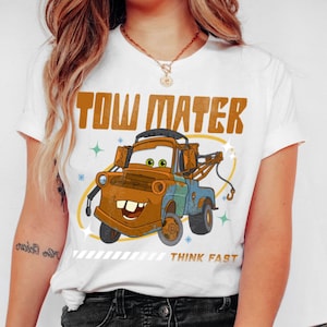 May include: A white short-sleeve t-shirt featuring a graphic of the rusty brown tow truck character Tow Mater. The text TOW MATER appears in large brown letters above the truck, with the words THINK FAST printed in smaller brown text below.