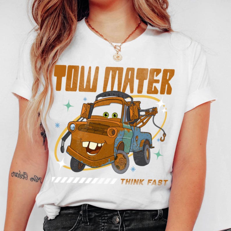 May include: A white short-sleeve t-shirt featuring a graphic of the rusty brown tow truck character Tow Mater. The text TOW MATER appears in large brown letters above the truck, with the words THINK FAST printed in smaller brown text below.