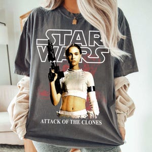 May include: Dark gray t-shirt featuring the Star Wars logo and a graphic of a woman holding a blaster. The text "ATTACK OF THE CLONES" is printed below the graphic. The shirt has short sleeves and a relaxed fit.