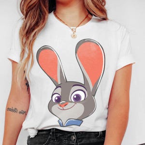 Puede incluir: Camiseta blanca con una imagen de dibujos animados de Judy Hopps de la pel&iacute;cula Zootr&oacute;polis. El gr&aacute;fico muestra la cara de Judy con pelaje gris, orejas internas rosas y ojos morados. La camiseta tiene mangas cortas y cuello redondo.