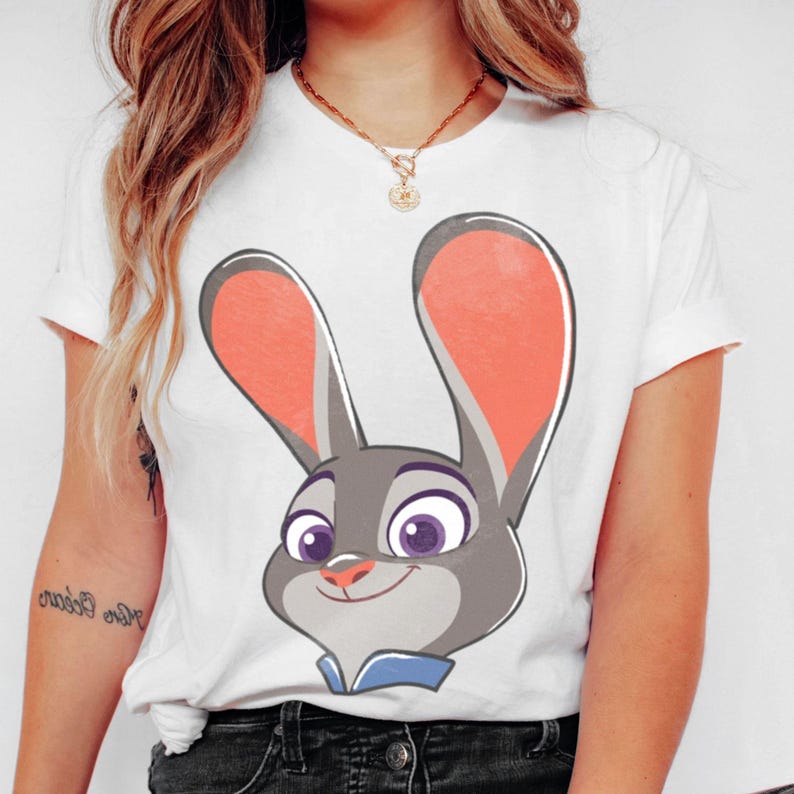 Puede incluir: Camiseta blanca con una imagen de dibujos animados de Judy Hopps de la pel&iacute;cula Zootr&oacute;polis. El gr&aacute;fico muestra la cara de Judy con pelaje gris, orejas internas rosas y ojos morados. La camiseta tiene mangas cortas y cuello redondo.