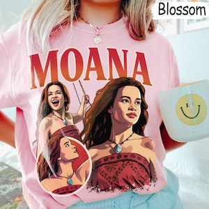 以下が含まれることがあります： 大きな赤い文字でMOANAと書かれたライトピンクのTシャツ。グラフィックには、赤い柄のトップスとネックレスを身につけたモアナのイラストが3つ描かれています。Tシャツはクルーネックで半袖です。