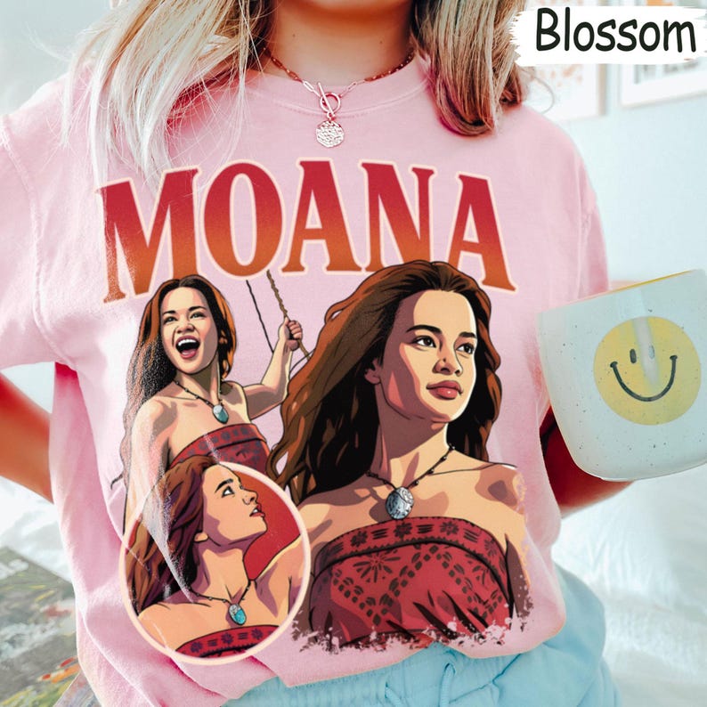 以下が含まれることがあります： 大きな赤い文字でMOANAと書かれたライトピンクのTシャツ。グラフィックには、赤い柄のトップスとネックレスを身につけたモアナのイラストが3つ描かれています。Tシャツはクルーネックで半袖です。