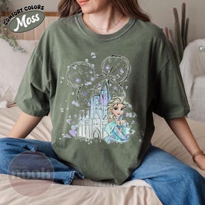 May include: Moss green t-shirt featuring a graphic of Elsa from Frozen, a castle, and Mickey Mouse ears. The design includes purple and blue accents. The shirt has the words "Comfort Colors" and "Moss" printed on the top left.