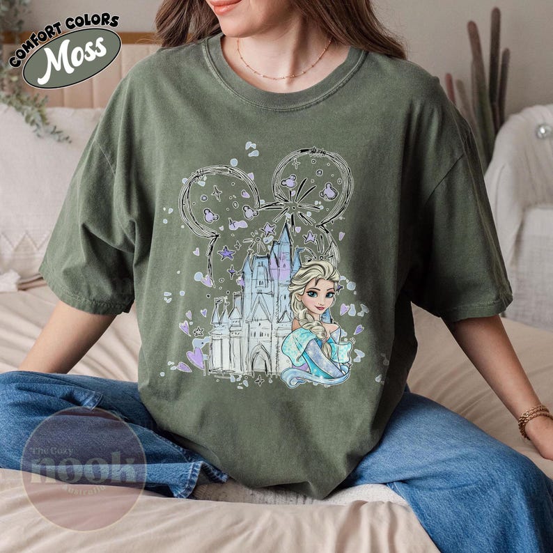 May include: Moss green t-shirt featuring a graphic of Elsa from Frozen, a castle, and Mickey Mouse ears. The design includes purple and blue accents. The shirt has the words "Comfort Colors" and "Moss" printed on the top left.