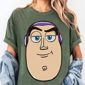 May include: Olive green t-shirt featuring a cartoon illustration of Buzz Lightyear's face. The graphic includes his head, face, and a portion of his shoulders. The character's face is tan with blue eyes, purple accents, and a black swirl.