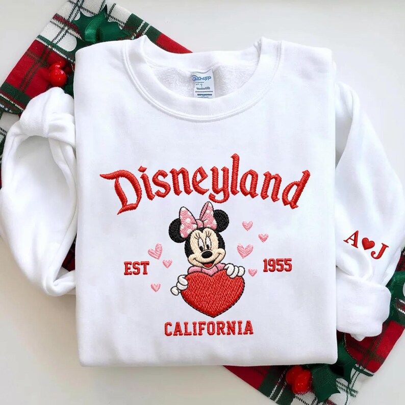 Puede incluir: Sudadera blanca con bordado rojo "Disneyland", Minnie Mouse con un coraz&oacute;n y "EST 1955 CALIFORNIA". La manga tiene las iniciales "A&hearts;J". La sudadera est&aacute; sobre un fondo a cuadros rojos y verdes.