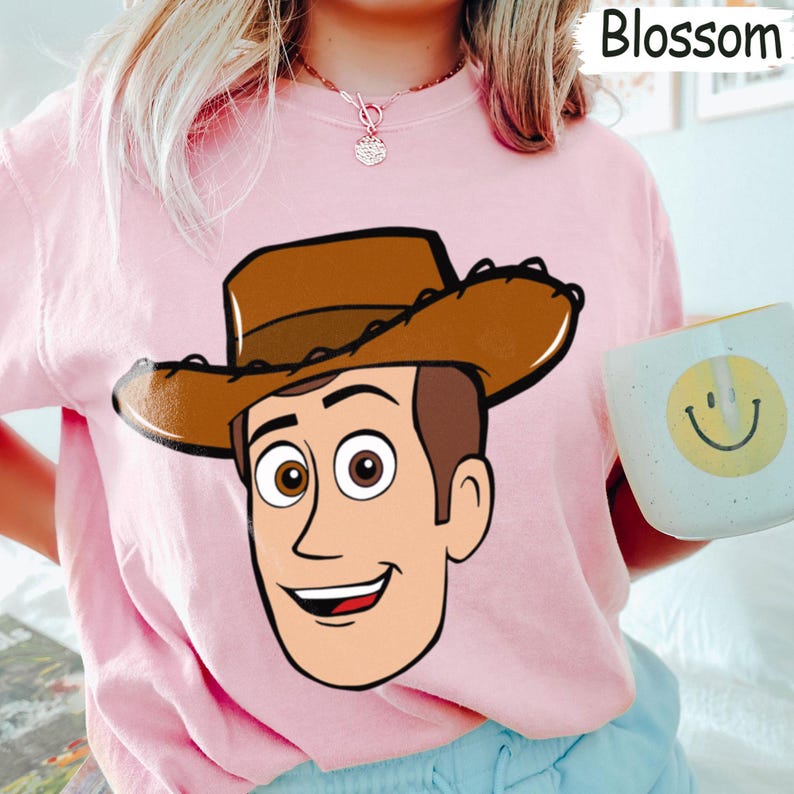May include: A light pink sweatshirt featuring a cartoon illustration of Woody from Toy Story. The character has brown hair, a cowboy hat, and a smiling expression. The word "Blossom" is in the top right corner. A white mug with a yellow smiley face is held in the right hand.