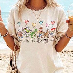 May include: Beige t-shirt with a cartoon graphic of Toy Story characters holding balloons. Characters include Mr. Potato Head, Buzz Lightyear, Rex, Woody, Bullseye, and Bo Peep. The text below the characters reads their names.