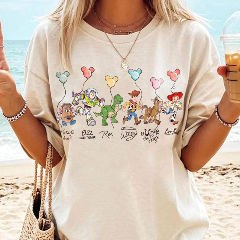 May include: Beige t-shirt with a cartoon graphic of Toy Story characters holding balloons. Characters include Mr. Potato Head, Buzz Lightyear, Rex, Woody, Bullseye, and Bo Peep. The text below the characters reads their names.