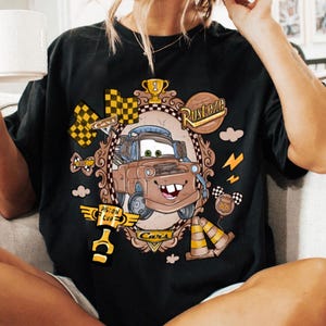May include: Black t-shirt featuring a cartoon image of Mater from the movie Cars, framed in an ornate design with the words "Rusteze" and "Piston Cup". Additional graphics include checkered flags and lightning bolts.