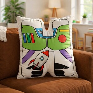 Puede incluir: Un coj&iacute;n decorativo blanco con un dise&ntilde;o de dibujos animados de un guardabosques espacial. El coj&iacute;n presenta un casco verde, detalles rojos y azules, brazos morados y un cohete. El coj&iacute;n est&aacute; sobre un sof&aacute; marr&oacute;n.