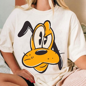 May include: Cream-colored t-shirt featuring a cartoon illustration of Pluto's face. The design showcases Pluto's yellow face, black ears, and expressive eyes, outlined in black. A classic and playful design.