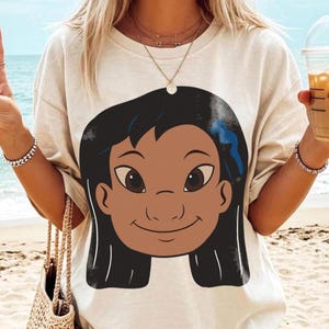 Puede incluir: Camiseta de color crema con una ilustraci&oacute;n de dibujos animados de una ni&ntilde;a sonriente con cabello negro y un lazo azul. El gr&aacute;fico tiene piel morena y ojos marrones grandes. La camiseta es de estilo casual, adecuada para el uso diario.