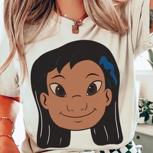 Puede incluir: Camiseta blanca con una ilustraci&oacute;n de dibujos animados de una ni&ntilde;a con cabello negro, piel morena y un accesorio azul para el cabello. El gr&aacute;fico est&aacute; centrado en la parte delantera de la camiseta.