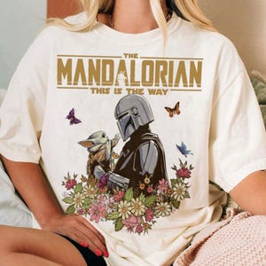 May include: Cream-colored t-shirt featuring a graphic of The Mandalorian and Grogu surrounded by flowers and butterflies. The text "THE MANDALORIAN" and "THIS IS THE WAY" are printed above the illustration. The shirt is a casual wear item.