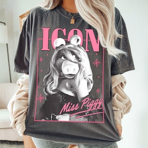 May include: Dark gray t-shirt featuring a graphic of Miss Piggy from The Muppets. The design includes the word "ICON" in pink block letters above Miss Piggy's image, and "Miss Piggy" in cursive below. The shirt has a vintage, worn look.