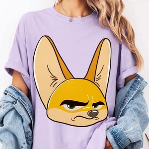 May include: A light purple t-shirt featuring a cartoon fox face with large ears and a grumpy expression. The fox has yellow and tan fur, green eyes, and a black nose. The shirt has short sleeves and a relaxed fit.