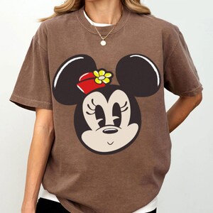 Peut inclure: T-shirt marron avec un motif de Minnie Mouse. Le motif montre le visage de Minnie avec des oreilles noires, un n&oelig;ud rouge et une fleur jaune. Le t-shirt a des manches courtes et un col rond.