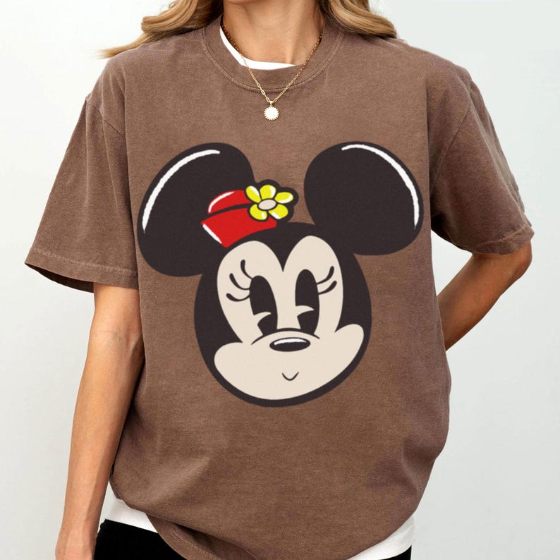 Peut inclure: T-shirt marron avec un motif de Minnie Mouse. Le motif montre le visage de Minnie avec des oreilles noires, un n&oelig;ud rouge et une fleur jaune. Le t-shirt a des manches courtes et un col rond.