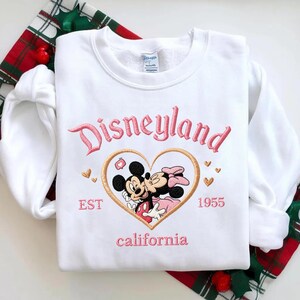 Peut inclure: Sweat-shirt blanc avec texte brod&eacute; rose "Disneyland" au-dessus d'un motif en forme de c&oelig;ur avec Mickey et Minnie Mouse. Le motif comprend le texte "EST 1955 California". Le sweat-shirt est sur un fond &agrave; carreaux rouges et verts.