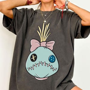 May include: Dark gray t-shirt featuring a cartoon voodoo doll design. The doll has a light blue face with a pink bow, yellow hair, and stitched details. The shirt has short sleeves and a relaxed fit.