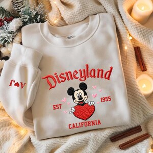 Puede incluir: Sudadera blanca con bordado rojo "Disneyland", Mickey Mouse sosteniendo un coraz&oacute;n rojo y "California" debajo. El pu&ntilde;o de la manga tiene iniciales rojas y un coraz&oacute;n. El dise&ntilde;o est&aacute; sobre una tela blanquecina.