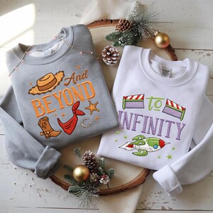 Puede incluir: Dos sudaderas con dise&ntilde;os bordados. Una es gris con "And Beyond" y gr&aacute;ficos tem&aacute;ticos de vaqueros. La otra es blanca con "To Infinity" y un dise&ntilde;o de cohete. Ambas son de cuello redondo.