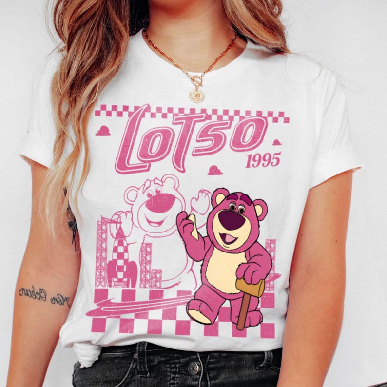 May include: White t-shirt featuring a pink graphic of Lotso, the bear from Toy Story, with the text "Lotso 1995". The design includes a checkered pattern and a rocket ship illustration. The shirt has short sleeves and a crew neck.
