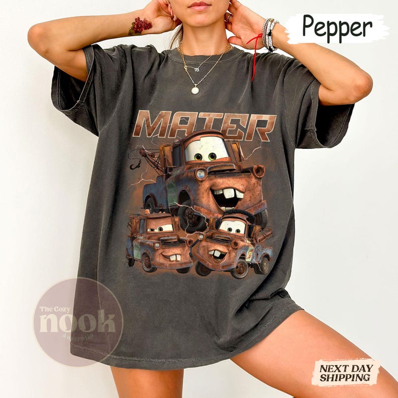 May include: Dark grey t-shirt with a graphic of Mater from the film Cars. The word "MATER" is printed above the character. The shirt has a relaxed fit and short sleeves. The Cozy Nook logo is visible.