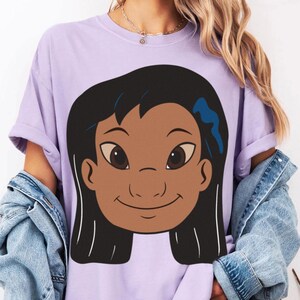 Puede incluir: Una camiseta de color lila con una ilustraci&oacute;n de dibujos animados de una ni&ntilde;a sonriente con cabello negro y ojos marrones. El gr&aacute;fico incluye un accesorio para el cabello azul. La camiseta tiene mangas enrolladas y se usa con una chaqueta vaquera.