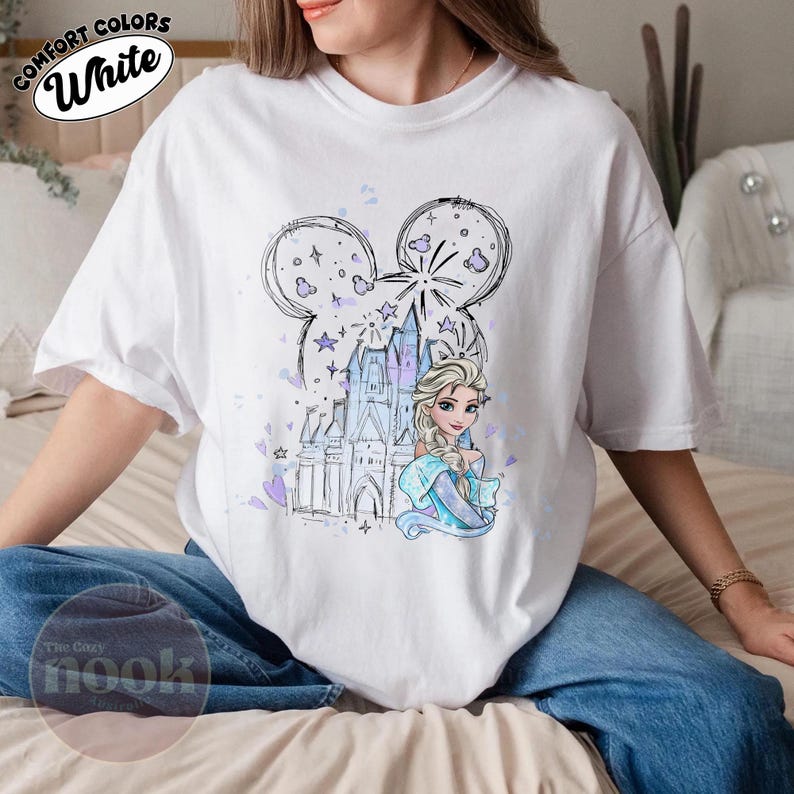 May include: White t-shirt featuring a cartoon illustration of Elsa from Frozen, a castle, and a Mickey Mouse head outline. The design includes purple and blue accents. The words "Comfort Colors" and "White" are printed on the shirt.