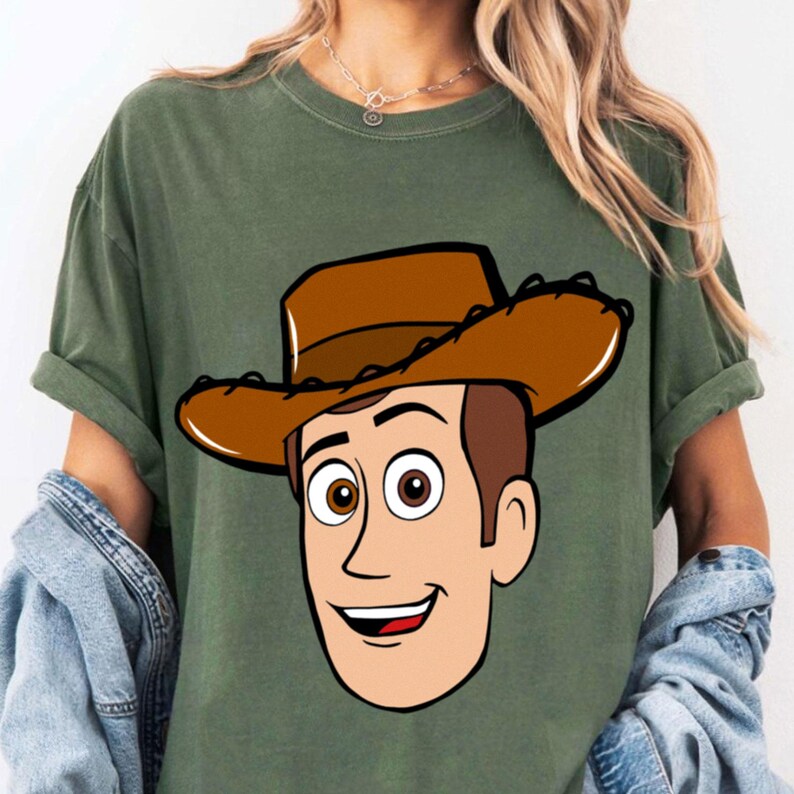 May include: Olive green t-shirt featuring a cartoon illustration of Woody from Toy Story. The graphic shows Woody's face, complete with a brown cowboy hat, brown hair, and a smiling expression. The shirt has rolled-up sleeves.