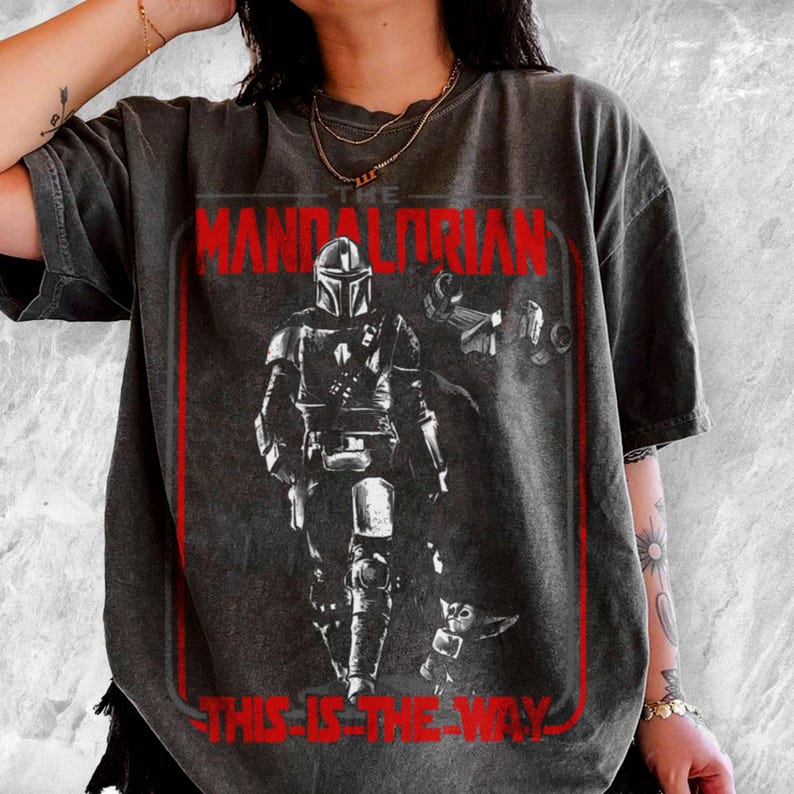 May include: Dark gray t-shirt featuring a red framed graphic of The Mandalorian and Grogu. The text "THE MANDALORIAN" and "THIS IS THE WAY" are in red. The shirt has a vintage, distressed look.