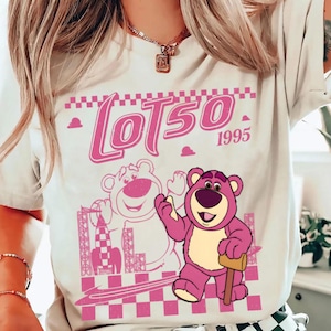 May include: A light-colored t-shirt featuring a pink graphic of Lotso, a bear character, with the text "Lotso 1995". The design includes a checkered pattern and a rocket ship illustration. The bear is holding a mallet.