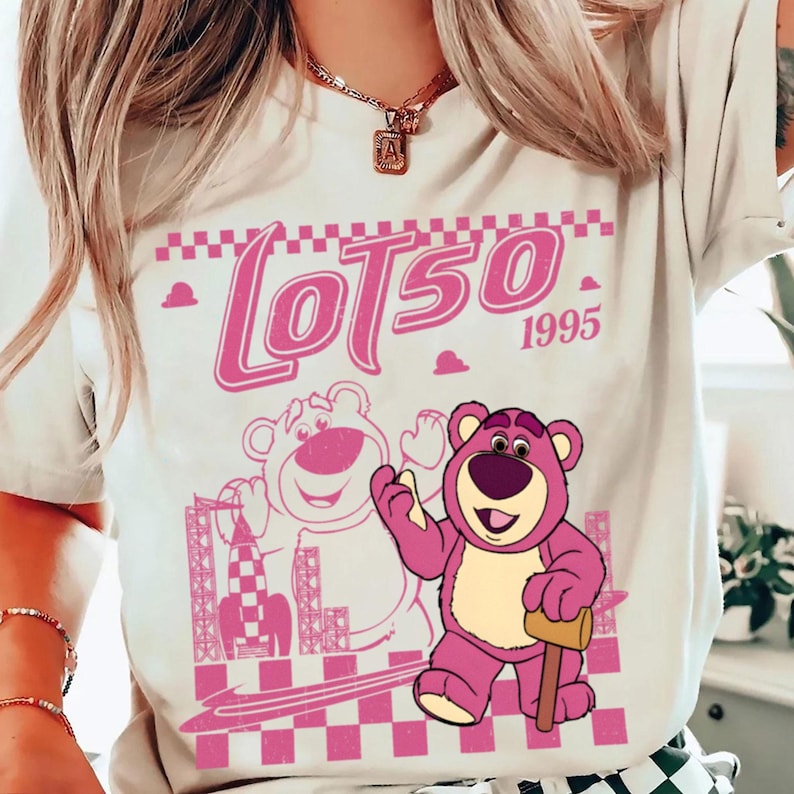 May include: A light-colored t-shirt featuring a pink graphic of Lotso, a bear character, with the text "Lotso 1995". The design includes a checkered pattern and a rocket ship illustration. The bear is holding a mallet.