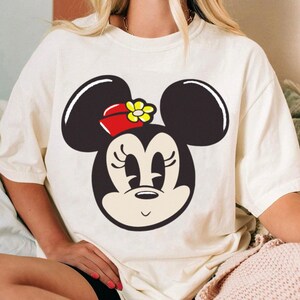 Peut inclure: T-shirt cr&egrave;me avec un grand dessin du visage de Minnie Mouse. Le personnage de dessin anim&eacute; a des oreilles noires, un n&oelig;ud rouge avec une fleur jaune et une expression souriante. Le t-shirt est de coupe ample.