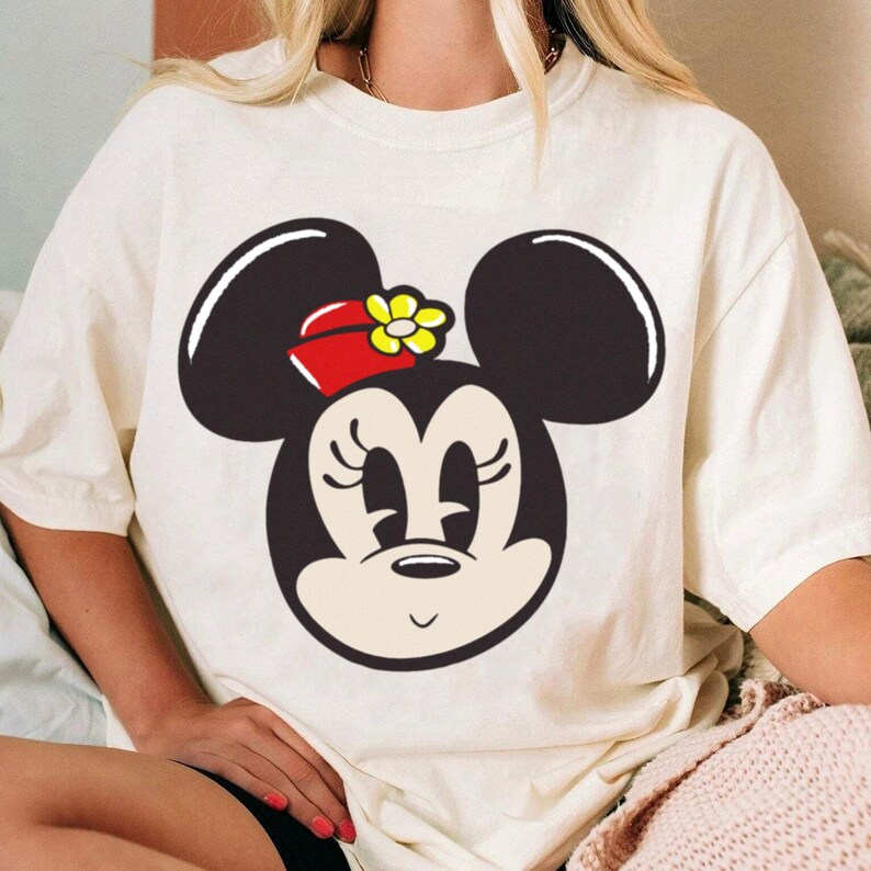 Peut inclure: T-shirt cr&egrave;me avec un grand dessin du visage de Minnie Mouse. Le personnage de dessin anim&eacute; a des oreilles noires, un n&oelig;ud rouge avec une fleur jaune et une expression souriante. Le t-shirt est de coupe ample.