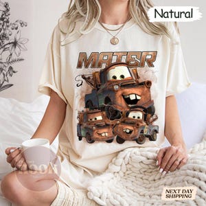 May include: Cream-coloured t-shirt featuring a graphic of the character Mater from the animated film Cars. The design includes the word "Mater" in a brown, distressed font, and images of Mater in various sizes. The shirt has a relaxed fit.